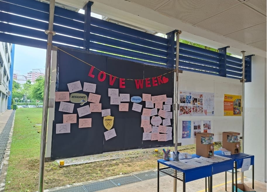 An outdoor noticeboard decorated with a "Love Week" banner displays pink cards organised under values including Resilience, Integrity, and Courage, written by students.